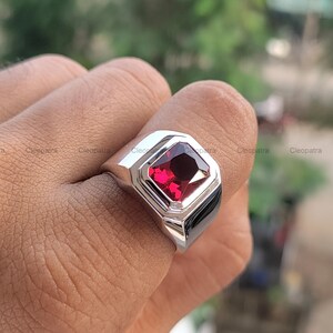 Puede incluir: Un anillo de plata con una gran piedra preciosa roja oscura de talla cuadrada. El anillo tiene un diseño geométrico moderno con un acabado pulido. La piedra preciosa está engastada en un bisel.