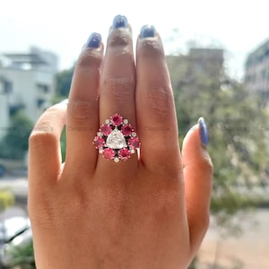 May include: A silver ring with a large triangular diamond surrounded by smaller red gemstones. The ring is set on a white band.