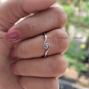 May include: A silver ring with a swirling design. The ring has a delicate, openwork spiral at the centre, with a slender band that curves around the finger. The ring is shown on a hand with pink nail polish.