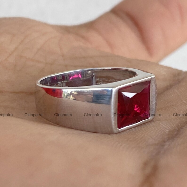 May include: A silver-toned ring featuring a prominent, square-cut, deep red gemstone set into a smooth, polished metal band. The design showcases a clean, geometric aesthetic with the vibrant red stone serving as the central focal point of the piece.