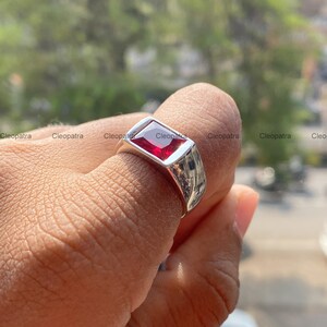 May include: A silver-toned ring featuring a rectangular, faceted red gemstone set in a smooth, polished metal band. The ring has a wide, solid shank that tapers slightly toward the bottom, showcasing a clean, reflective surface and a vibrant red stone.