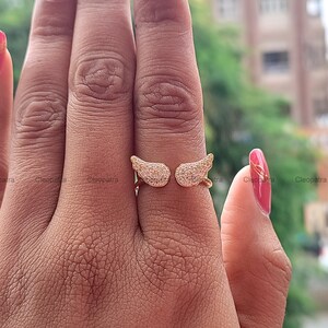 May include: A gold-colored ring featuring an open design with two wing-shaped elements. Each wing is encrusted with small, sparkling stones, creating a shimmering effect. The ring is shown on a hand with a pink manicure.