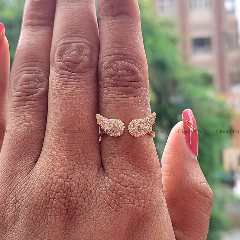 May include: A gold-colored ring featuring an open design with two wing-shaped elements. Each wing is encrusted with small, sparkling stones, creating a shimmering effect. The ring is shown on a hand with a pink manicure.