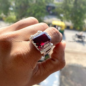 Pode incluir: Um grande anel de prata ornamentado com uma pedra preciosa retangular vermelho escuro como peça central. O anel tem um design detalhado e texturizado com pedras retangulares claras menores em ambos os lados da pedra principal.