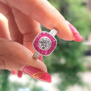 Puede incluir: Primer plano de un anillo plateado con una piedra central grande, redonda y transparente. La piedra central está rodeada por un halo de pequeñas piedras rosas cuadradas. La banda del anillo también está adornada con pequeñas piedras transparentes.
