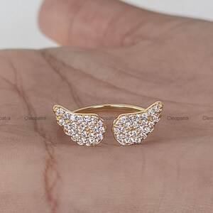 May include: A gold-colored ring featuring two openwork wing designs, each encrusted with small, clear gemstones. The ring is set against a neutral background, highlighting the sparkle of the stones.