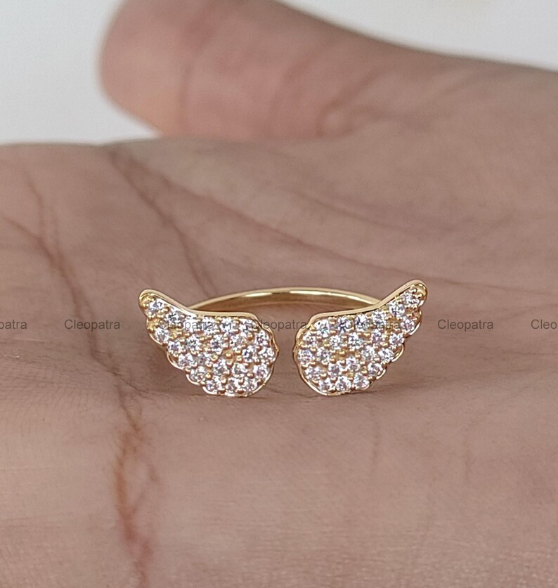 May include: A gold-colored ring featuring two openwork wing designs, each encrusted with small, clear gemstones. The ring is set against a neutral background, highlighting the sparkle of the stones.