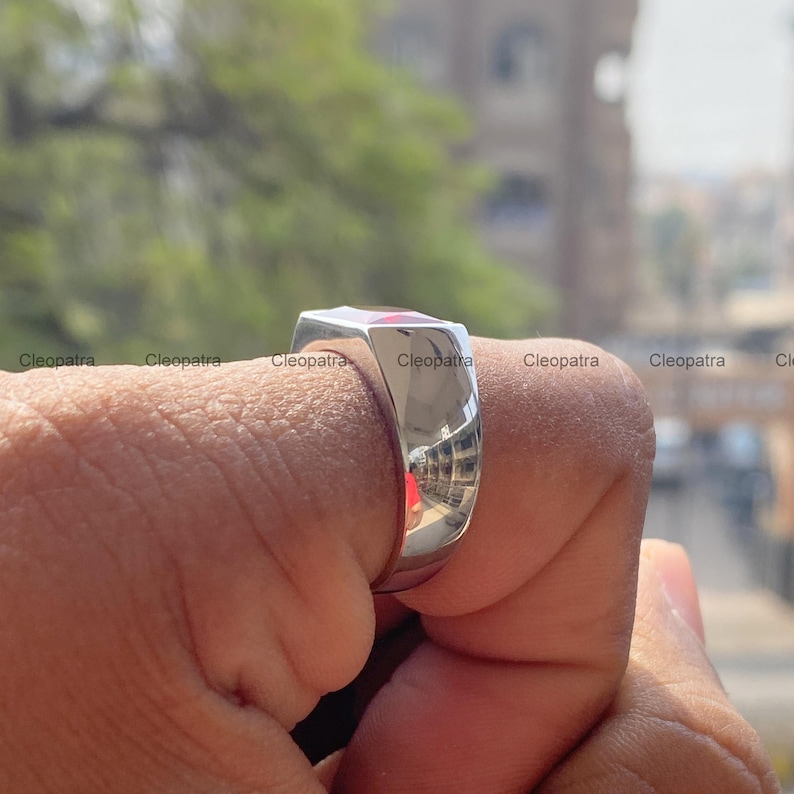 May include: A polished silver-tone signet ring with a smooth, reflective surface and a flat top. The metal band features a clean, solid construction with a high-shine finish that captures light and surrounding reflections on its curved exterior.