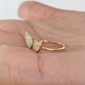 May include: A gold-colored ring with a butterfly wing design. The wings are encrusted with small, clear gemstones. The ring is shown on a person's palm.