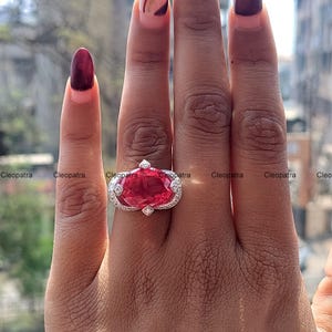 Puede incluir: Un anillo de plata con una piedra preciosa roja grande de forma ovalada rodeada de piedras preciosas más pequeñas y transparentes.