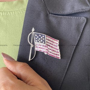 Pode incluir: Um broche de bandeira em tom prateado com um campo azul retangular com pequenos detalhes de estrelas e listras horizontais compostas por fileiras alternadas de pedras vermelhas e transparentes de corte baguette em uma haste de metal torcido.