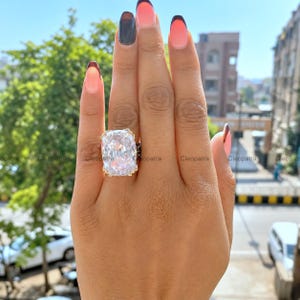 Peut inclure: Une grande bague ornée d'une pierre précieuse transparente taille coussin, montée sur un anneau en métal doré. La bague présente une pierre centrale facettée proéminente avec un éclat brillant, maintenue par de petites griffes.