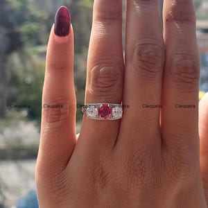 Peut inclure: Une bague en argent avec une pierre précieuse rouge et deux petites pierres blanches. La bague est portée sur le doigt d'une personne.