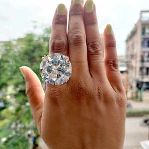 Könnte beinhalten: Ein großer, runder, klarer Edelsteinring mit einem vielseitigen Design. Der Ring ist auf einem silbernen Band mit vier Krappen gefasst. Die Hand trägt gelben Nagellack.