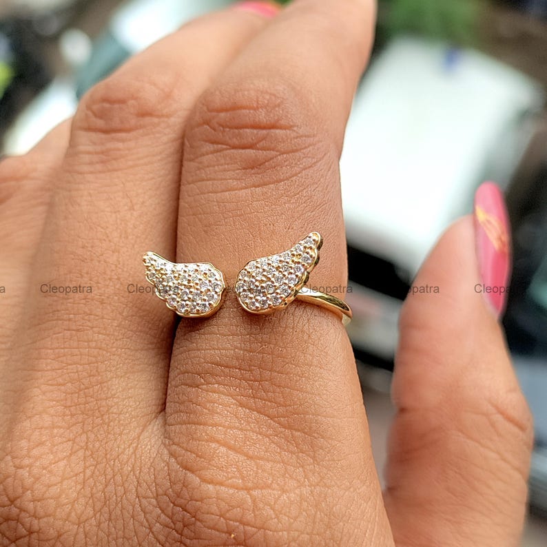 May include: A gold-colored ring featuring an open design with two wing-shaped elements. Each wing is encrusted with small, clear gemstones, creating a sparkling effect. The ring is designed to wrap around the finger, offering a unique and stylish look.