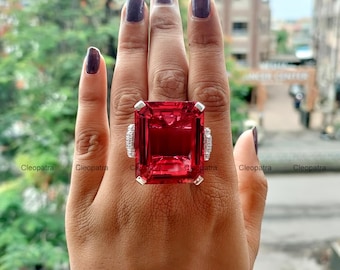 May include: A large, rectangular, ruby-red gemstone ring with a silver band. The ring has small, white accent stones on each side of the main stone. The ring is displayed on a hand.