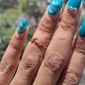 May include: A gold-colored ring spelling out the word "fuck" in a cursive font. The ring is on a finger with turquoise and white marbled nail polish. The word "Cleopatra" is printed in white on the image.