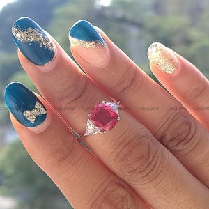 May include: A close-up of a hand wearing a silver ring with a large red gemstone and smaller white gemstones. The hand is also wearing teal blue nail polish with gold glitter accents.