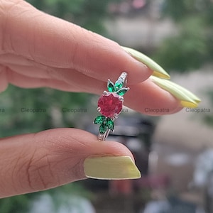 May include: A silver ring featuring a large, round, red gemstone, accented with green leaf-shaped stones. The ring has a delicate band with small, clear stones. The ring is held up by a person's fingers.
