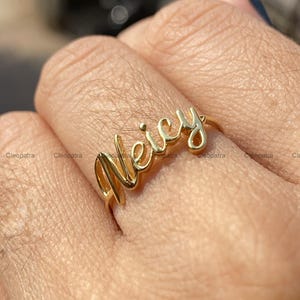 Könnte beinhalten: Ein goldfarbener Ring mit dem Namen "Weicy" in Schreibschrift. Der Ring wird an einem Finger getragen und zeigt das filigrane Design und den personalisierten Schmuck. Der Ring ist ein individuelles Schmuckstück.