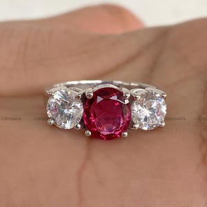 May include: A silver ring with a central oval ruby-red gemstone and two clear, round gemstones either side. The ring has a silver band. The gemstones are set in a three-stone design.