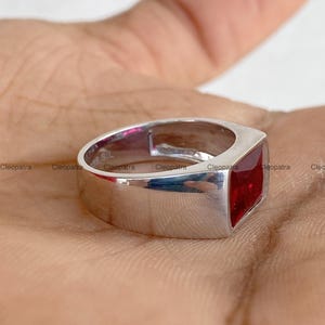 May include: A silver-toned ring featuring a square-cut, deep red gemstone set into a wide, polished band. The interior of the band is clearly marked with the inscription 925 SIL, indicating the metal composition of this piece of jewelry.