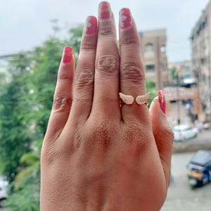 May include: A gold-colored ring with two open wings, each encrusted with small, clear stones. The ring is worn on a finger, showcasing the delicate design and sparkle. The ring is a jewelry item.