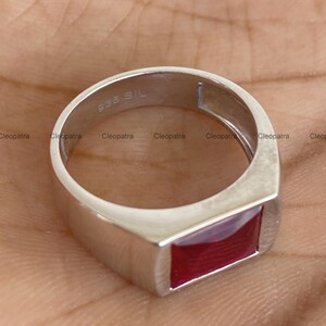 May include: A silver-toned ring featuring a rectangular, deep red gemstone set into the band. The interior of the band is clearly stamped with the text 935 SIL, indicating the metal composition. The design is smooth, polished, and metallic.