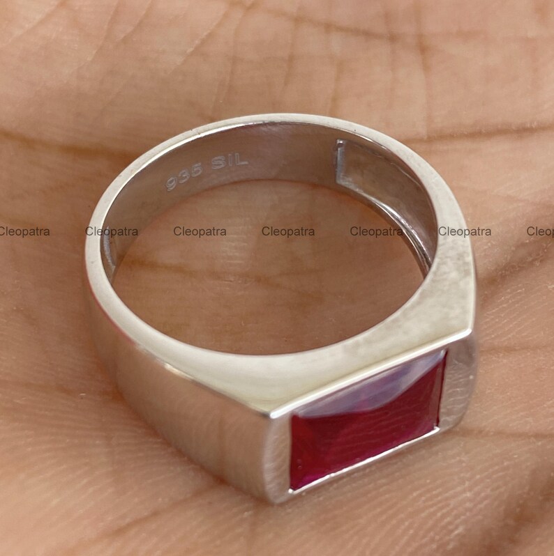 May include: A silver-toned ring featuring a rectangular, deep red gemstone set into the band. The interior of the band is clearly stamped with the text 935 SIL, indicating the metal composition. The design is smooth, polished, and metallic.