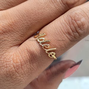 Pode incluir: Um anel dourado com o nome "Adele" escrito em itálico. O anel é usado em um dedo, com o nome curvando-se na parte superior. O anel é feito de um metal brilhante e reflexivo.