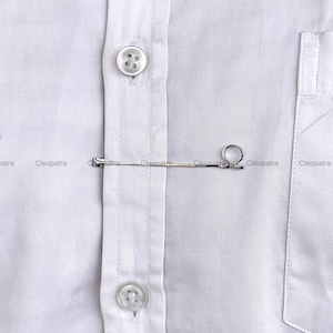 May include: A silver tie bar clipped onto a white shirt. The tie bar is a thin, straight piece of metal with a small loop at one end. The shirt has a button-down front and a pocket.