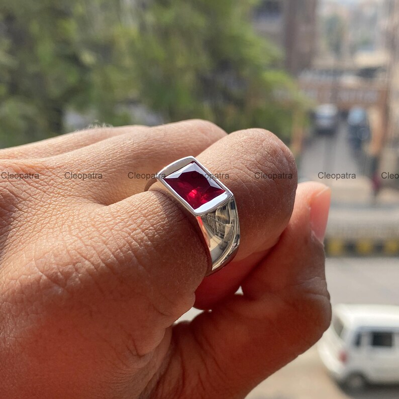 May include: A silver-toned ring featuring a rectangular, faceted, deep red gemstone set in a smooth, polished metal band. The ring has a wide, solid shank that tapers slightly toward the bottom, showcasing a reflective, metallic finish.