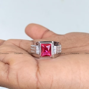 Puede incluir: Un anillo plateado con una gema roja rectangular vibrante en el centro. El anillo tiene un diseño moderno con pequeñas piedras claras de acento a cada lado de la piedra principal. El anillo se muestra en una mano.