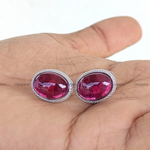 Può includere: Un paio di orecchini ovali con un vivace centro di pietra preziosa magenta, circondato da un alone di piccole pietre bianche scintillanti. Gli orecchini sono incastonati in un metallo color argento.