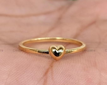 May include: A gold-colored ring featuring a small, heart-shaped design. The ring has a thin band and a polished finish, reflecting light. The heart is centered on the band, creating a simple yet elegant look. Jewelry.