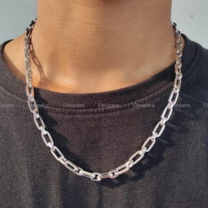 以下が含まれることがあります： 長方形のリンクデザインのシルバーチェーンネックレス。 ネックレスは首の周りに着用され、黒いTシャツの上に垂れ下がっています。 チェーンは光沢のある反射性の金属で作られています。