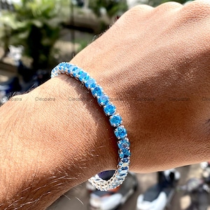 May include: A tennis bracelet featuring a series of round, bright blue gemstones set in a silver-colored metal. The bracelet is worn on a wrist, showcasing the vibrant color and design. The bracelet is a piece of jewelry.