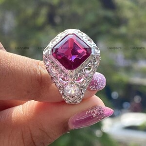 Puede incluir: Un anillo de tono plateado con una gran piedra preciosa magenta de corte cuadrado en el centro, rodeada de intrincados trabajos de metal en filigrana. Una piedra redonda transparente más pequeña está engastada bajo la gema principal.