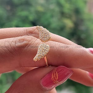 May include: A gold-colored ring with a unique design featuring two openwork wing shapes, each encrusted with small, sparkling stones. The ring is shown on a finger, highlighting its delicate details and craftsmanship.