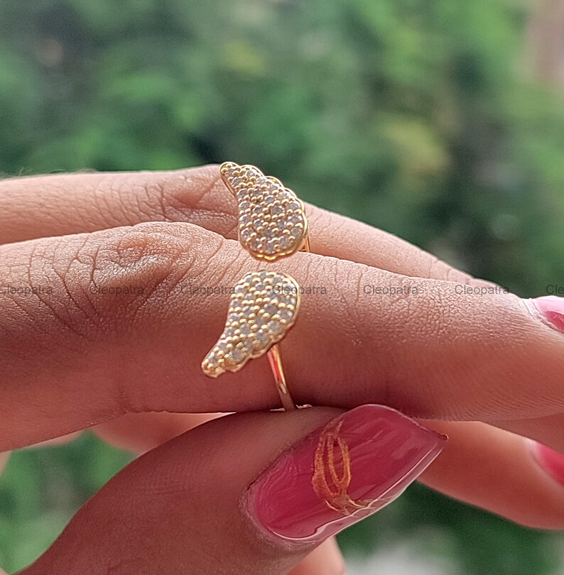 May include: A gold-colored ring with a unique design featuring two openwork wing shapes, each encrusted with small, sparkling stones. The ring is shown on a finger, highlighting its delicate details and craftsmanship.