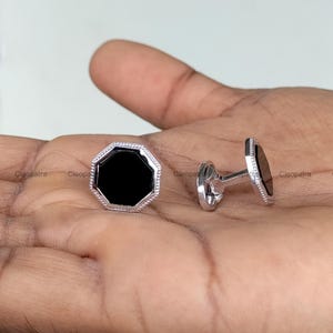 Puede incluir: Gemelos plateados con un centro de ónix negro octogonal. Los gemelos tienen un borde decorativo y un cierre de poste. La palabra "Cleopatra" está impresa en la imagen.