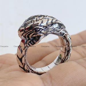 May include: A silver-toned metal ring featuring a detailed, sculpted dragon head design. The band of the ring is textured to resemble overlapping dragon scales that wrap around the finger, with a polished finish and darkened recessed areas.