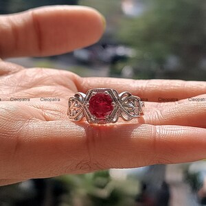 Könnte beinhalten: Ein silberner Ring mit einem roten Edelstein, der in einer sechseckigen Fassung sitzt. Der Ring hat ein filigranes, kunstvolles Design mit floralen Motiven.