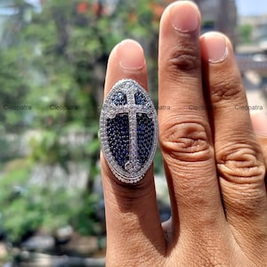 Puede incluir: Un anillo de plata con un diseño de cruz azul y blanco. El anillo es de forma ovalada y tiene una cruz en relieve en el centro. La cruz está rodeada de pequeñas piedras azules y el anillo está engastado con piedras blancas.