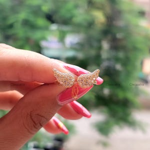May include: A pair of gold-colored angel wing earrings, encrusted with small, sparkling stones. The earrings are held up against a blurred green background, showcasing their intricate design and delicate details.