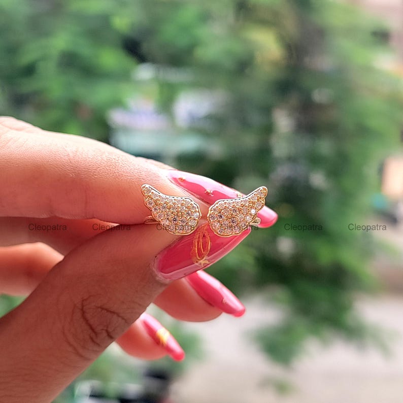 May include: A pair of gold-colored angel wing earrings, encrusted with small, sparkling stones. The earrings are held up against a blurred green background, showcasing their intricate design and delicate details.