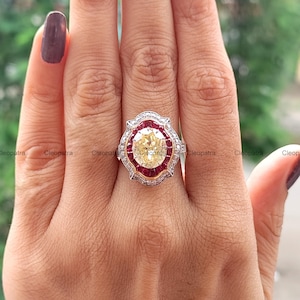 May include: A ring featuring a large oval-cut yellow centre stone surrounded by a halo of small red stones. The setting is framed by a scalloped border of tiny white stones, with a metallic band that appears to be a mix of silver and gold tones.
