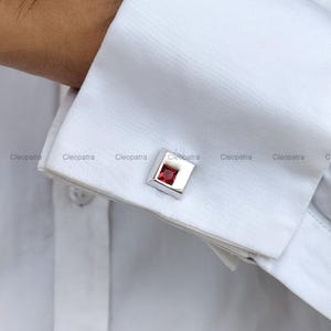 Puede incluir: Gemelos cuadrados plateados con una vibrante gema roja cuadrada en el centro. Los gemelos se muestran en un puño de camisa blanca. La palabra "Cleopatra" está impresa en la camisa.
