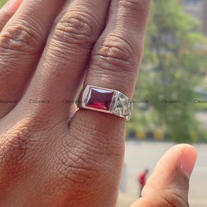 May include: A silver-toned metal ring featuring a rectangular, deep red gemstone set in the center. The band has a smooth, polished finish and a wide, flat top design that holds the faceted stone securely in place on the finger.