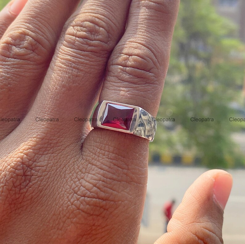 May include: A silver-toned metal ring featuring a rectangular, deep red gemstone set in the center. The band has a smooth, polished finish and a wide, flat top design that holds the faceted stone securely in place on the finger.